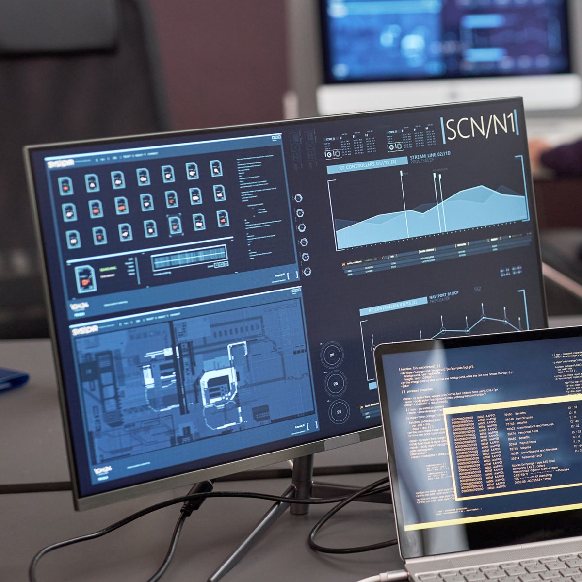 A workstation setup with multiple large monitors and a laptop displays dashboards, charts, and code, indicating real-time data analysis and system monitoring in an office setting, with a person working in the blurred background.