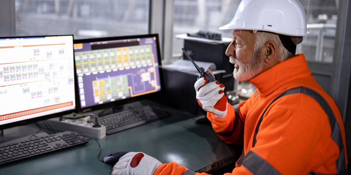 Person wearing a hard hat, gloves, and high-visibility clothing using a two-way radio beside computer monitors displaying system diagrams.