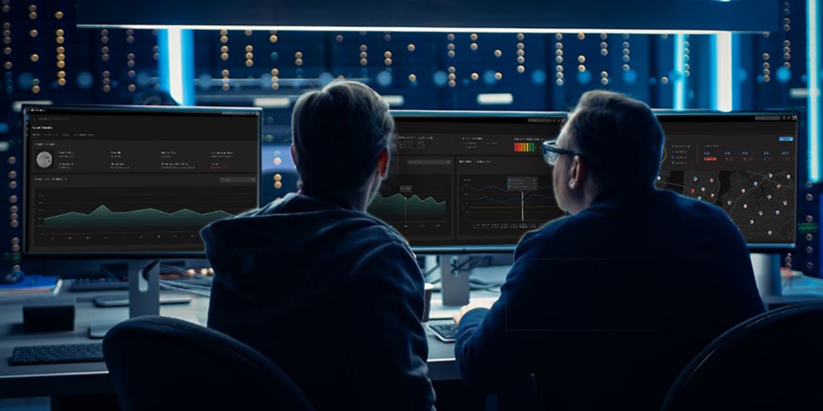 Two people seated at a workstation viewing multiple monitors displaying charts, graphs, maps, and dashboard interfaces in a technology environment.