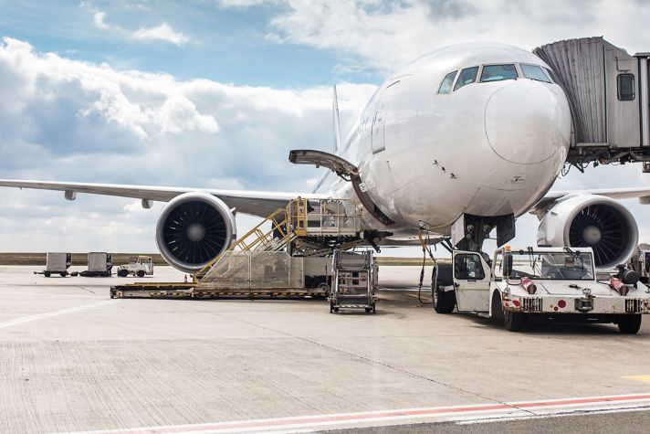 A large airplane is parked at the terminal, with ground crew loading cargo and preparing for departure under a cloudy sky.