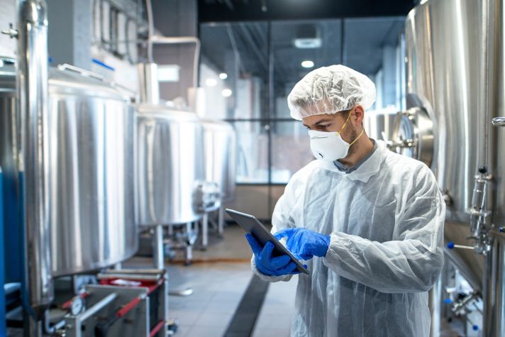 A technician wearing a lab coat and gloves uses a tablet to monitor processes in a modern brewing facility, surrounded by large metal brewing tanks.