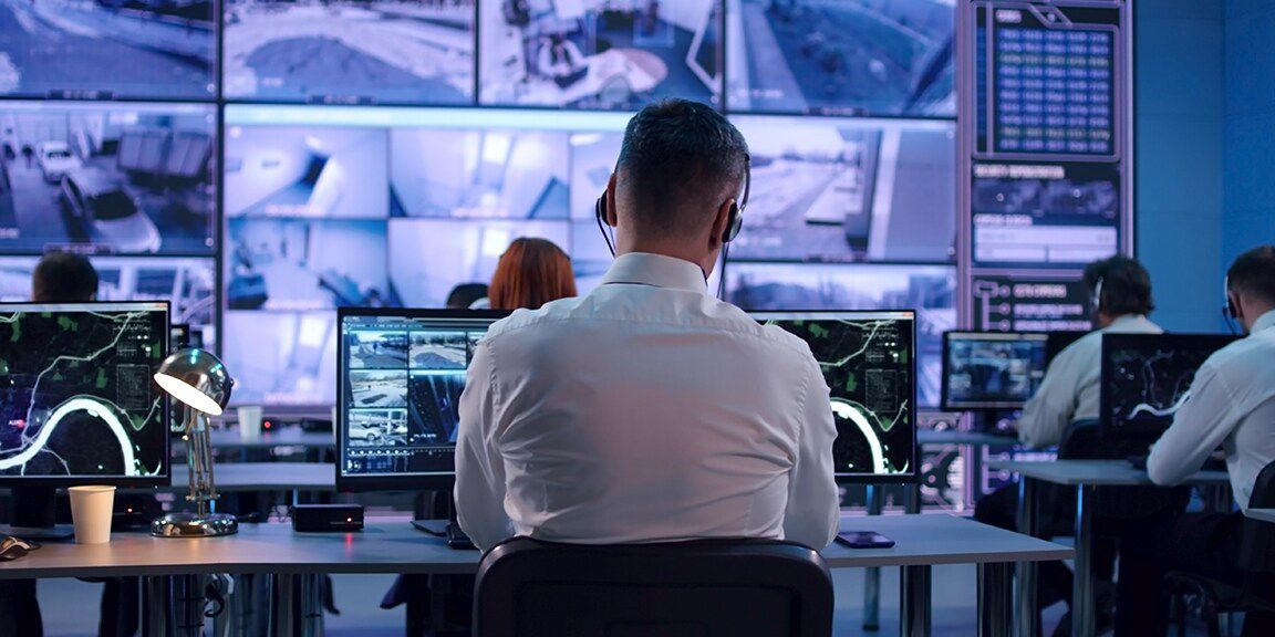 Analysts in a security monitoring room observe multiple screens displaying surveillance footage.