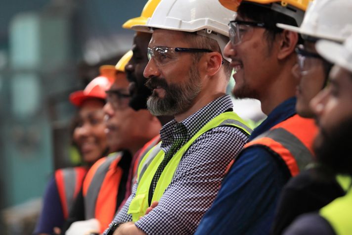 A group of workers in safety helmets and reflective vests, engaged in a discussion in an industrial setting, showcasing teamwork and safety protocols.