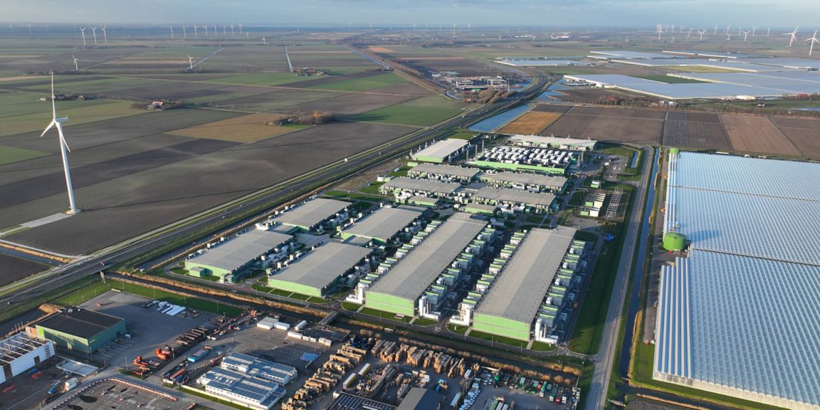 Aerial perspective of a green technology facility beside farmland, featuring wind turbines, solar panels, and industrial buildings in a rural landscape.