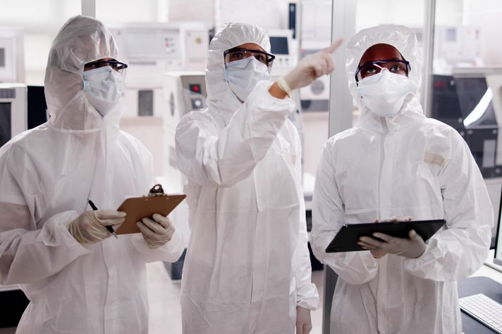 A group of three researchers wearing white protective suits and gloves collaborate in a laboratory setting. One researcher is holding a clipboard while another points toward a screen displaying data, indicating a technical discussion. The background features laboratory equipment, illustrating a high-tech scientific environment.