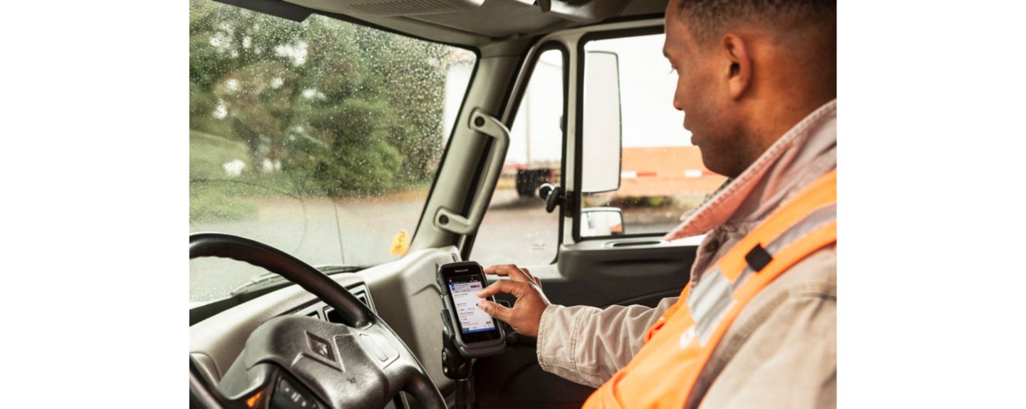 A person in an orange safety vest sits in a vehicle’s driver seat, checking a smartphone. The dashboard and steering wheel are clearly visible.