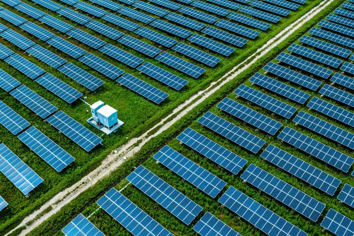Aerial view of a vast solar panel farm featuring rows of blue solar panels set on green grass. In the foreground, a small blue utility building is visible next to a dirt pathway that runs through the farm, providing access to the organized arrangement of solar panels.