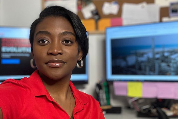 Chemical engineer Malaika Matumbu pictured in a red shirt in front of her workspace with two computer monitors.