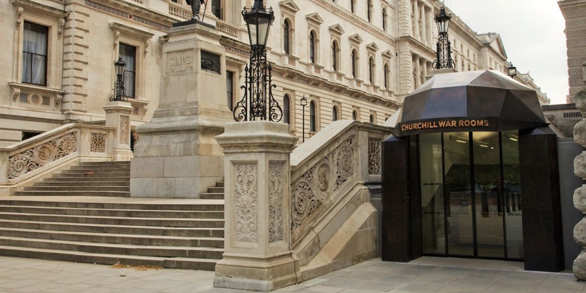 The entrance to the Churchill War Rooms, featuring a modern glass and metal structure with "CHURCHILL WAR ROOMS" inscribed above the door, complemented by ornate stone staircases and historical architecture in the background..