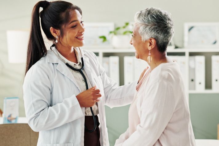 Healthcare professional in a white coat using a stethoscope while speaking with a seated patient in a modern clinic office with medical charts and shelves in the background.