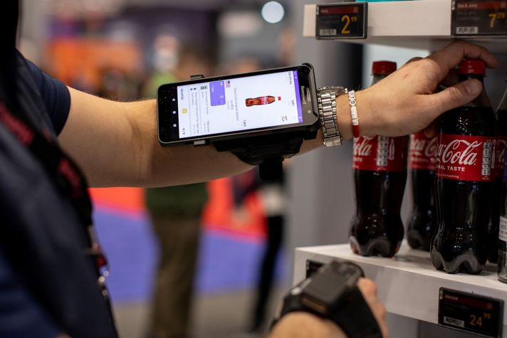 A person using a smartphone mounted on their arm to scan a Coca-Cola bottle from a shelf while managing inventory at a store. The phone screen displays product details and pricing information.