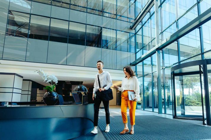 Man and woman walking in business lobby with a wall of windows behind them.