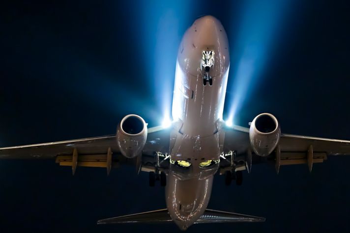 A commercial airplane is shown from below at night, with landing gear extended and bright lights illuminating its undercarriage and engines. The dark sky contrasts with the aircraft’s lighting, highlighting its structure and motion.