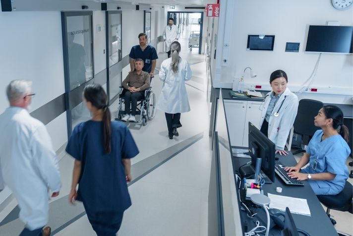 A busy hospital corridor featuring medical staff in white coats and scrubs attending to patients. A patient in a wheelchair is being escorted, with additional healthcare professionals walking and others at a reception desk. The environment portrays a professional healthcare setting.
