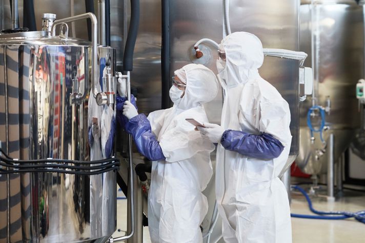 Two laboratory technicians in white protective suits are inspecting a large metallic tank. One uses a tool, while the other holds a smartphone. The background features industrial machinery and piping.