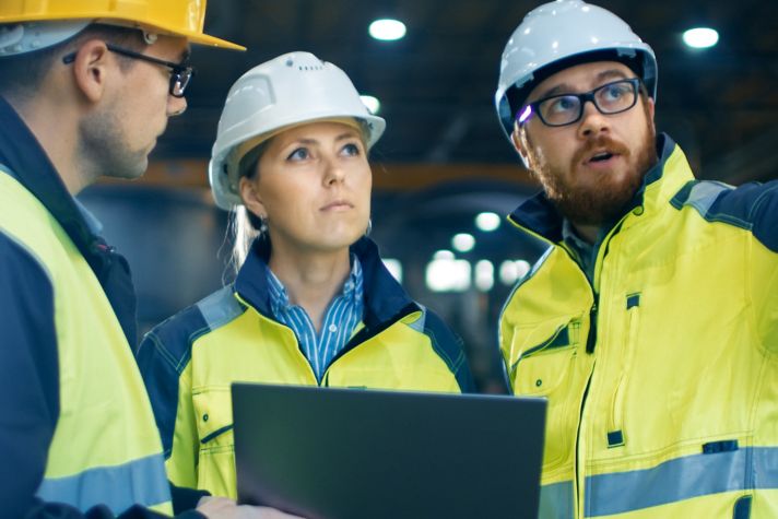 Three engineers wearing safety helmets and high-visibility jackets discussing a project inside an industrial facility. One engineer is holding a laptop, while another points towards something in the distance.