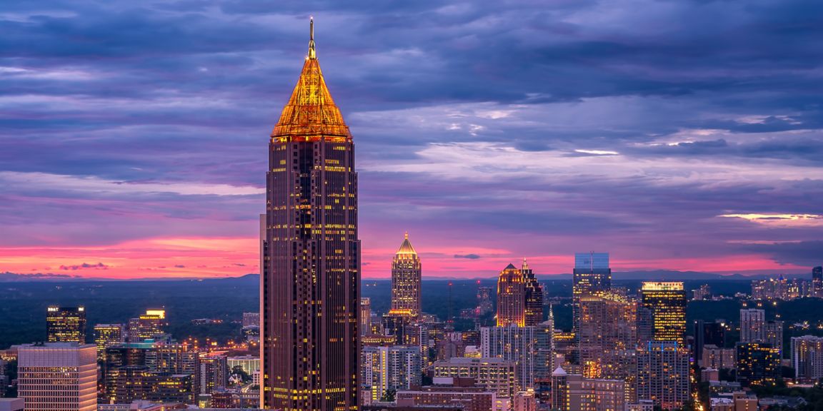A vibrant cityscape captured at dusk, featuring a tall building with a glowing pointed top. The sky is painted in shades of purple, pink, and blue, with clouds adding depth to the urban skyline.
