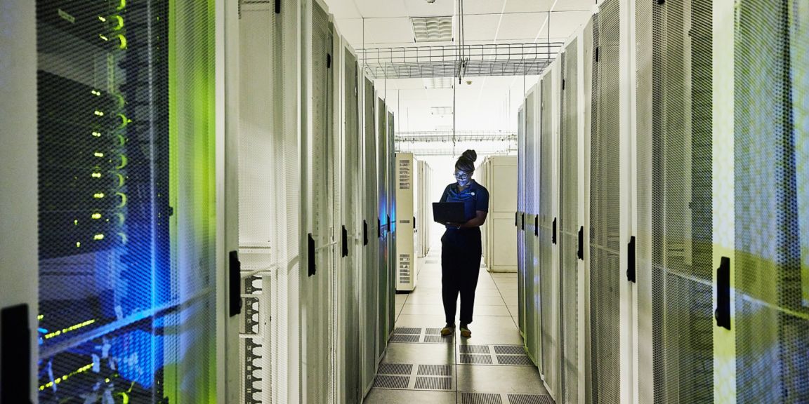 A professional works in a data center, surrounded by server racks while using a laptop. Soft blue and green lighting creates a high-tech atmosphere.
