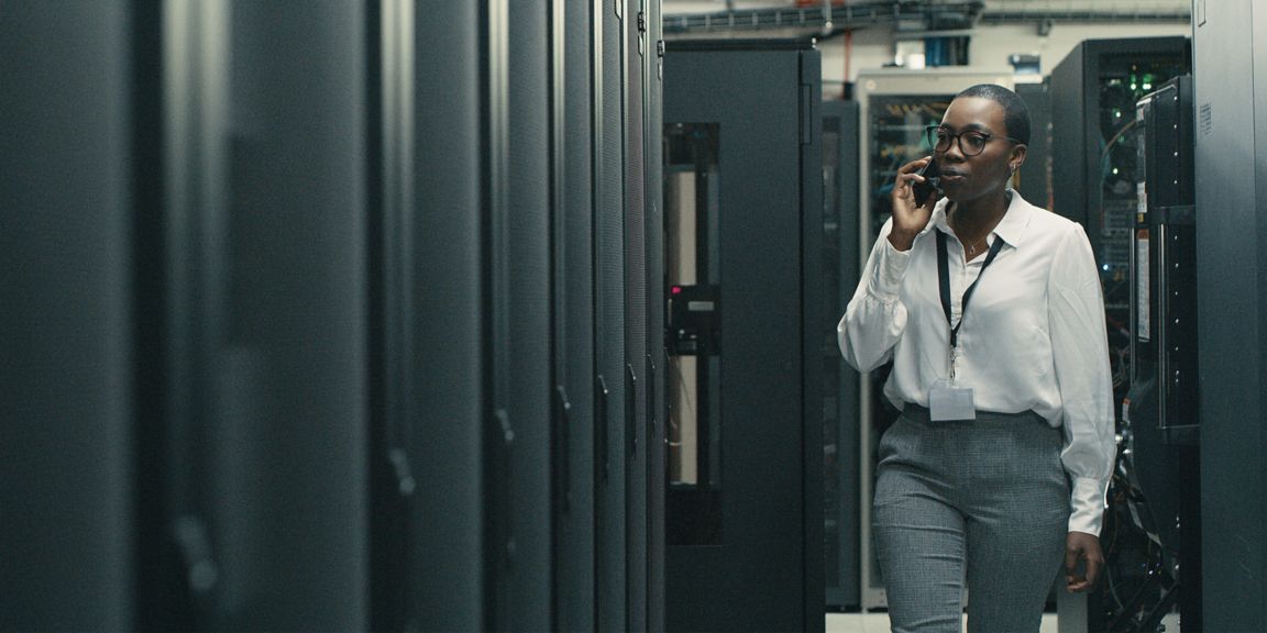 A person in business attire walks through a data center, engaged in a phone conversation, surrounded by rows of server racks.