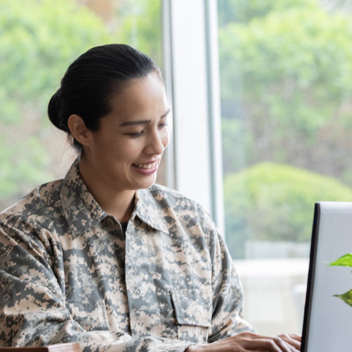 Soldiers in camouflage uniforms using laptops indoors near large windows.