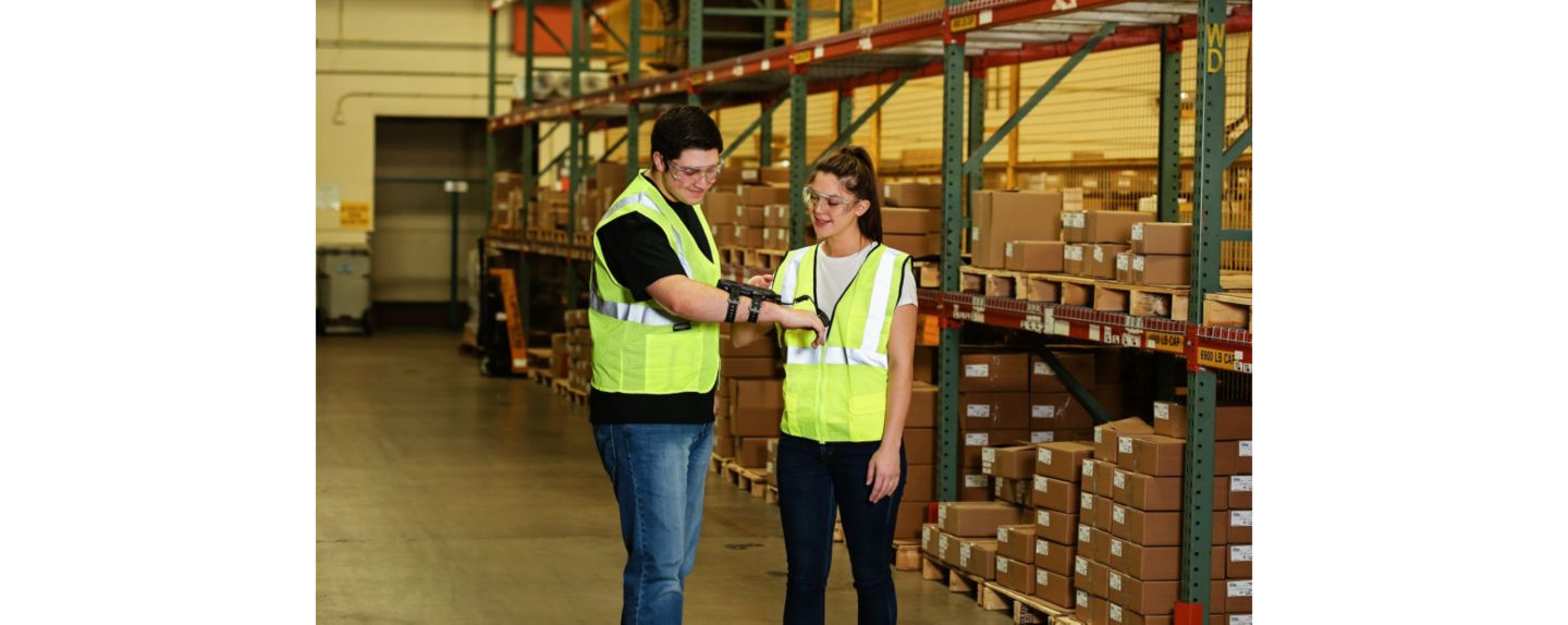 Two workers in high-visibility vests collaborate in a warehouse filled with shelves and boxes, emphasizing teamwork and operational efficiency.
