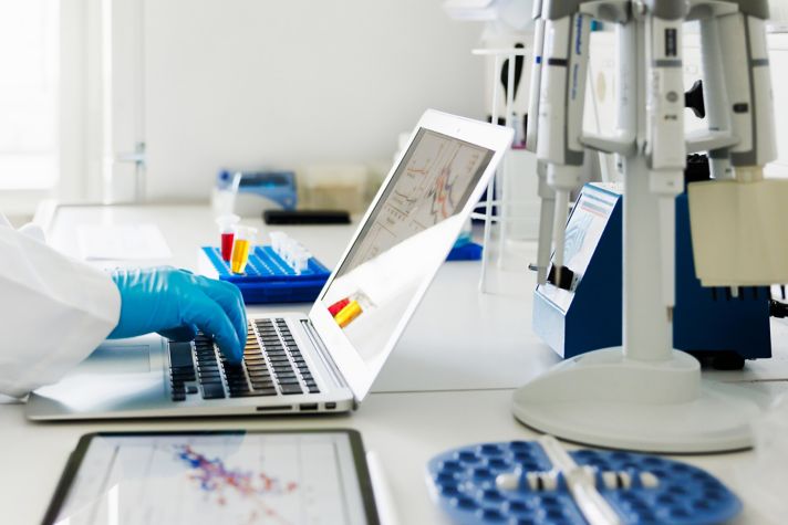 Scientist wearing blue gloves working on a laptop in a laboratory, surrounded by test tubes, a tablet displaying data charts, and other scientific instruments.
