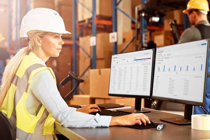 A person in a hard hat and safety vest works at a computer with two monitors, analyzing data in a warehouse environment filled with boxes.