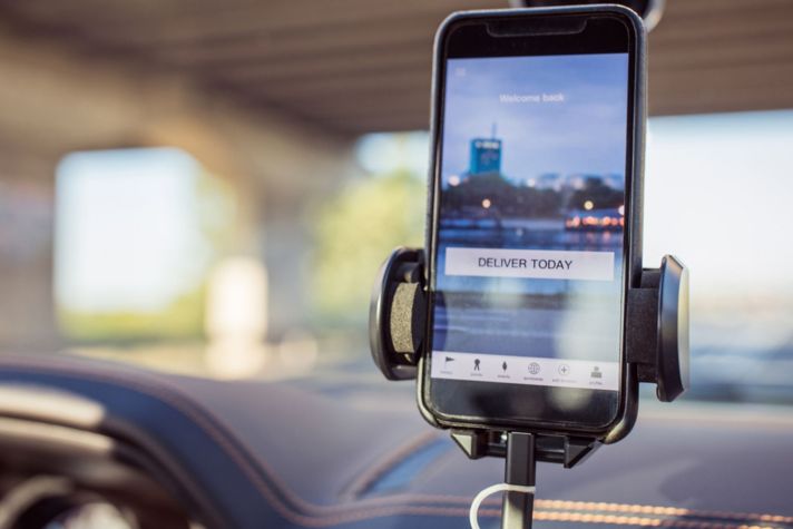 A phone screen shows a city background with the words "Deliver today" in a call-to-action button; the phone is positioned on a mounting device in a car.