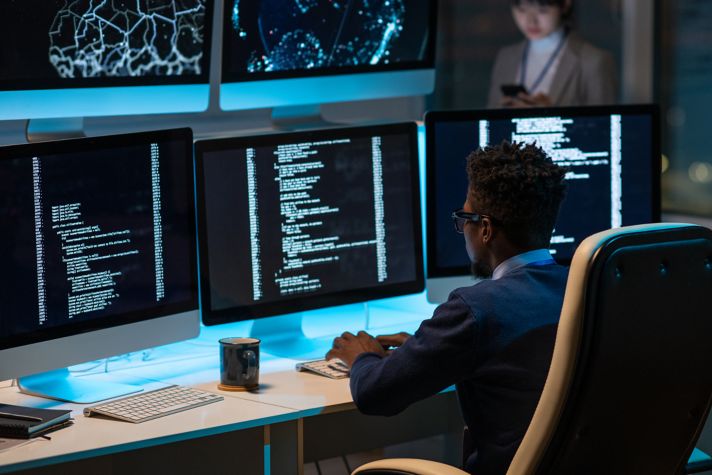 A software developer working at a desk with multiple monitors displaying code and data visualizations in a modern office setting. Another person is seen in the background using a smartphone.