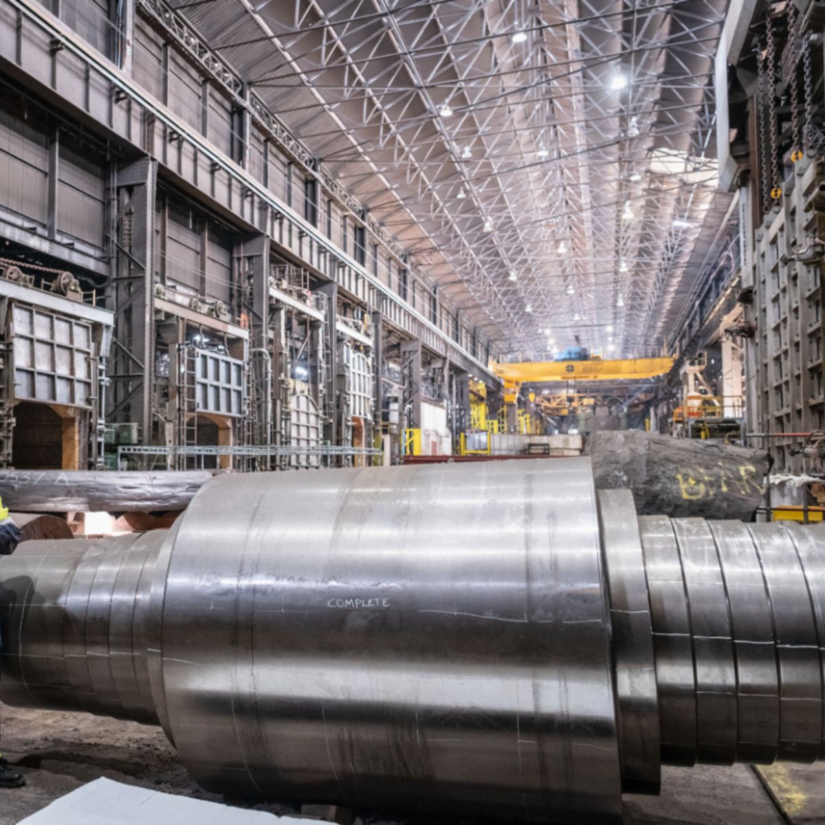 Two workers stand beside a large metal cylinder in an industrial facility with heavy machinery and overhead structures.