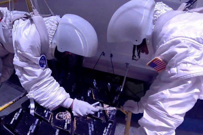 Two astronauts in white suits examine black samples in a space simulation environment, focusing on precise measurements for research purposes.