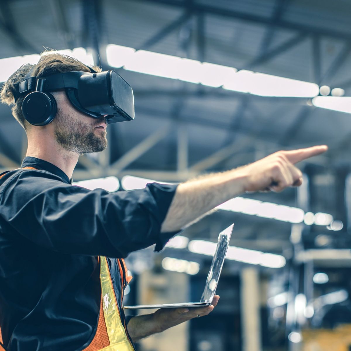 A man wearing a VR headset and headphones points while holding a laptop in a warehouse environment.