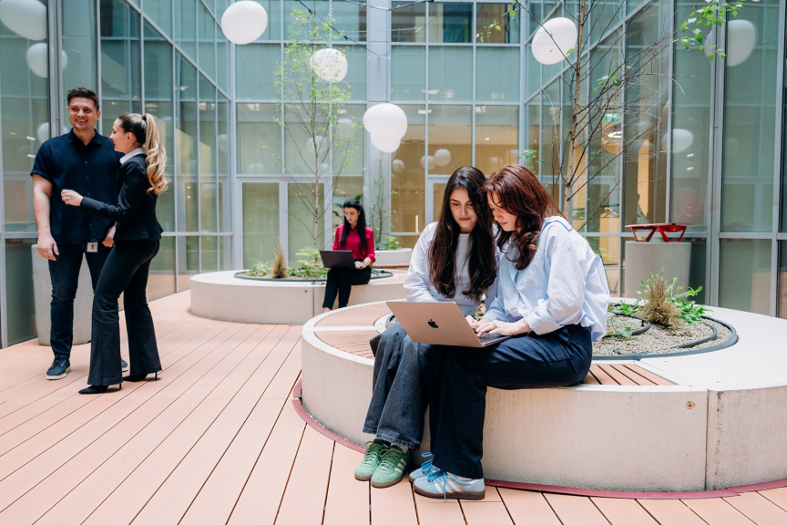 Diverse professionals work on laptops and converse within a modern, sunlit architectural atrium featuring natural elements and clean geometric design.
