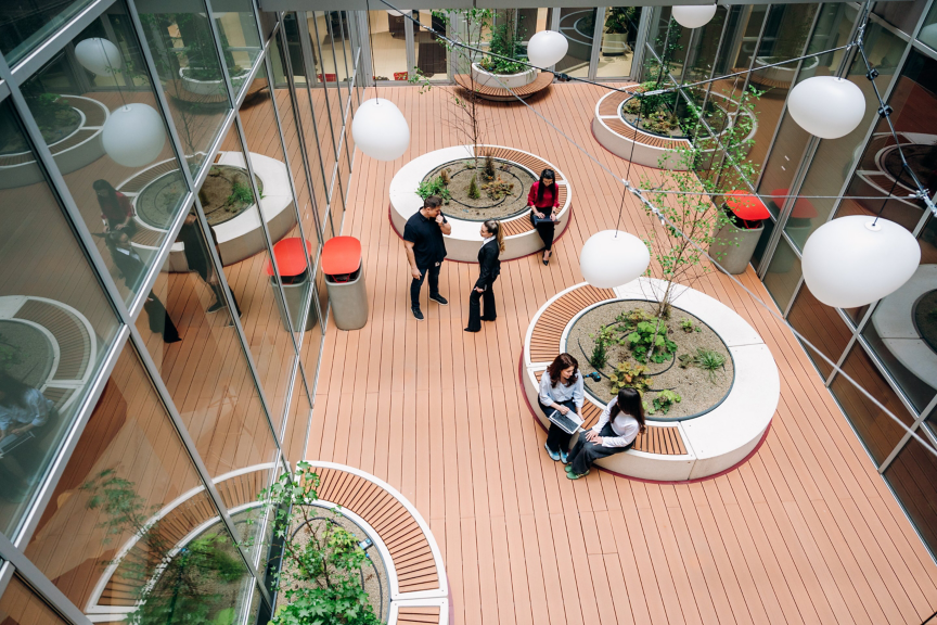A high-angle view of business professionals collaborating in a modern, landscaped office atrium with circular seating areas and glass-paneled walls.