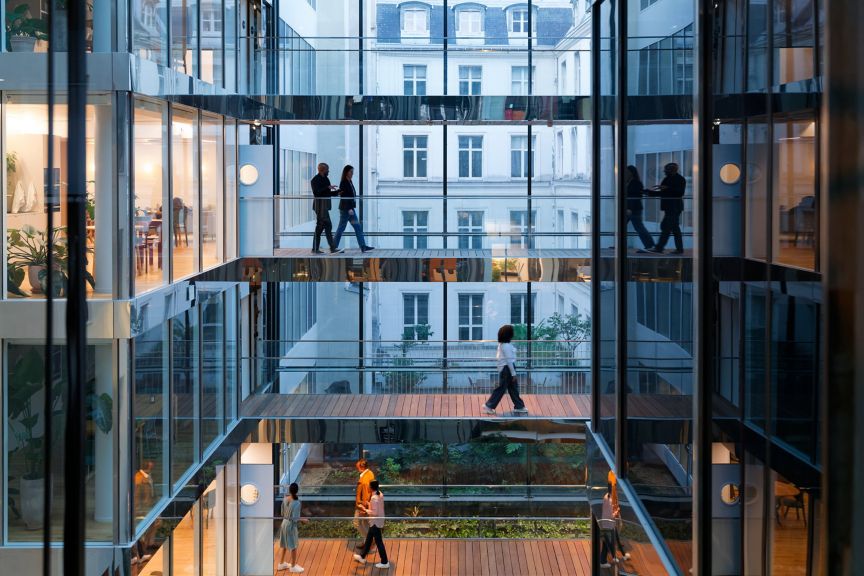 Employees navigate multi-level glass walkways in a modern office atrium featuring internal greenery and reflective architecture.