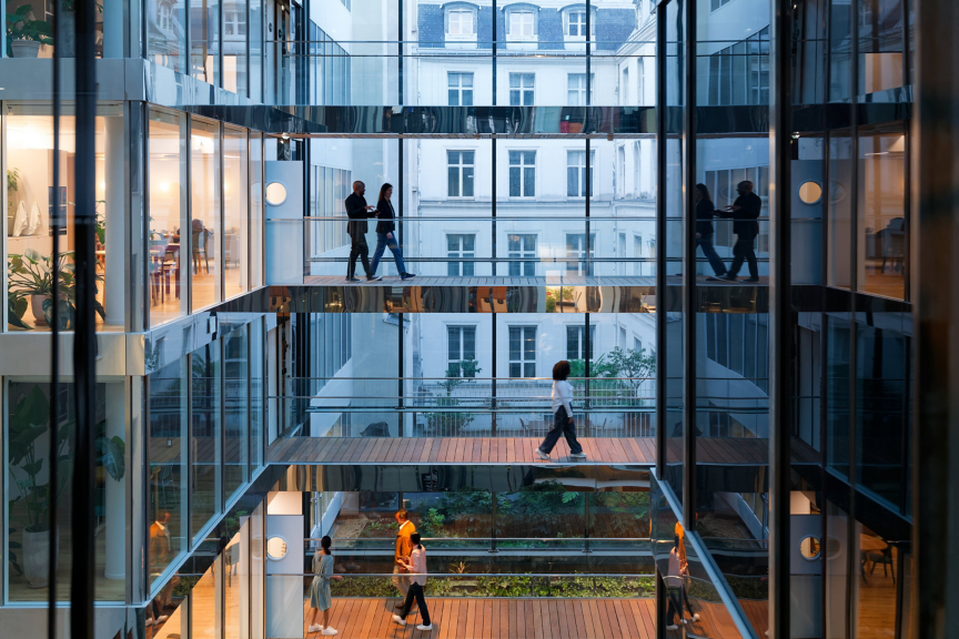 Employees navigate multi-level glass walkways in a modern office atrium featuring internal greenery and reflective architecture.
