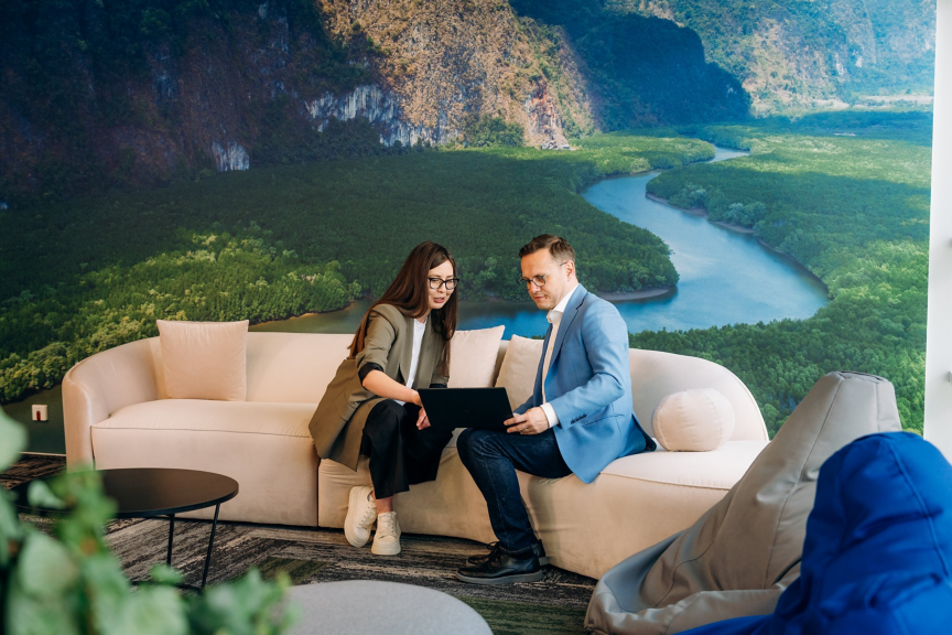 Two business professionals discuss data on a laptop while seated on a sofa against a large nature-themed mural in a modern office lounge area.