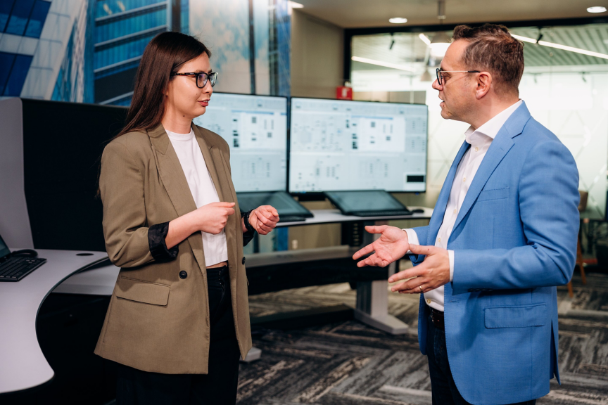Two professionals in business attire engage in a collaborative discussion in front of digital control room displays.
