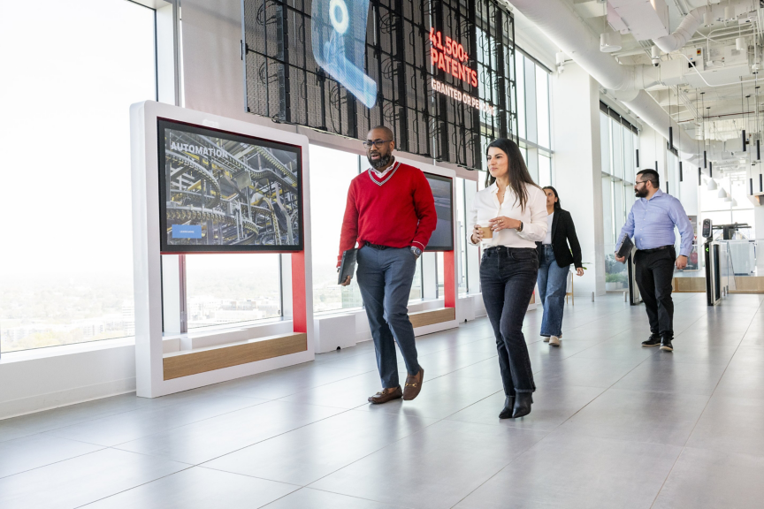 Diverse corporate team members walk through a modern, light-filled office hallway past digital displays showcasing company technology and achievements.
