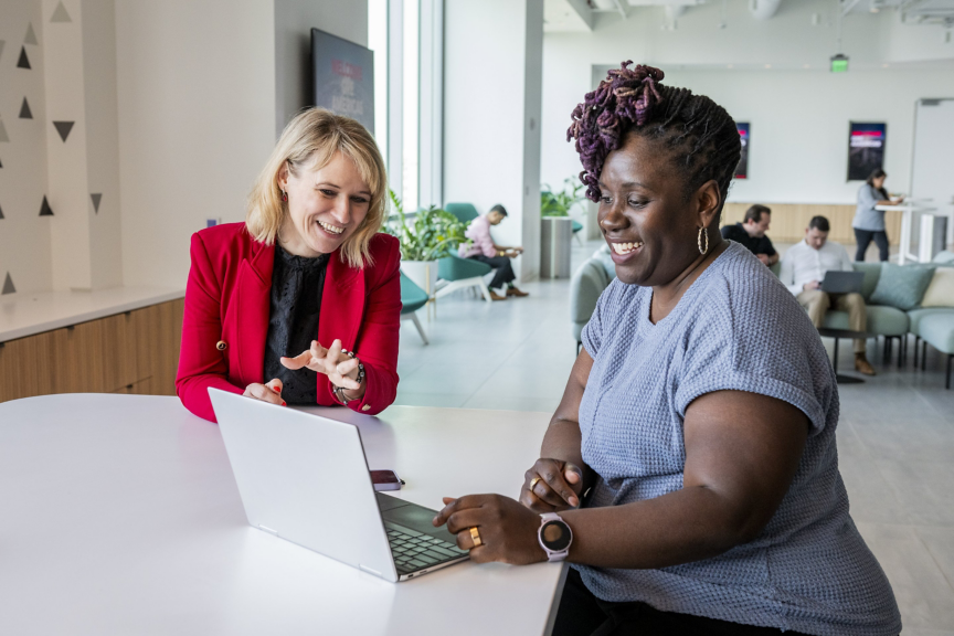 Two diverse professional women share a lighthearted, collaborative moment while working together on a laptop in a bright, modern office workspace.