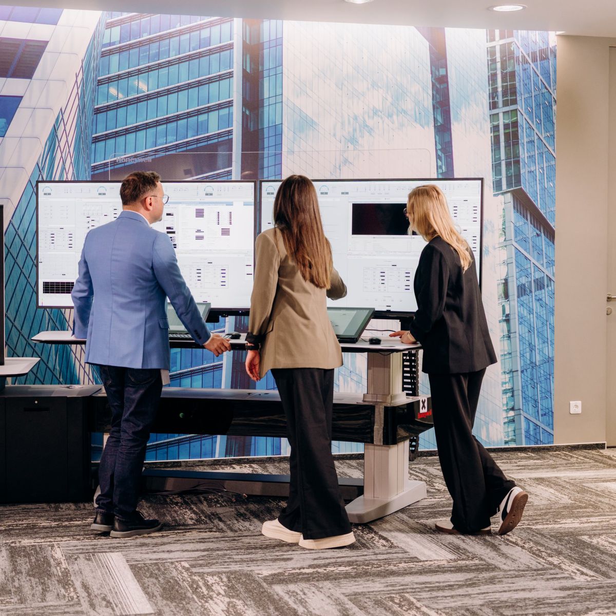 Three professionals in business attire collaborate while reviewing industrial metrics on large monitors in a modern, office-based control room environment.