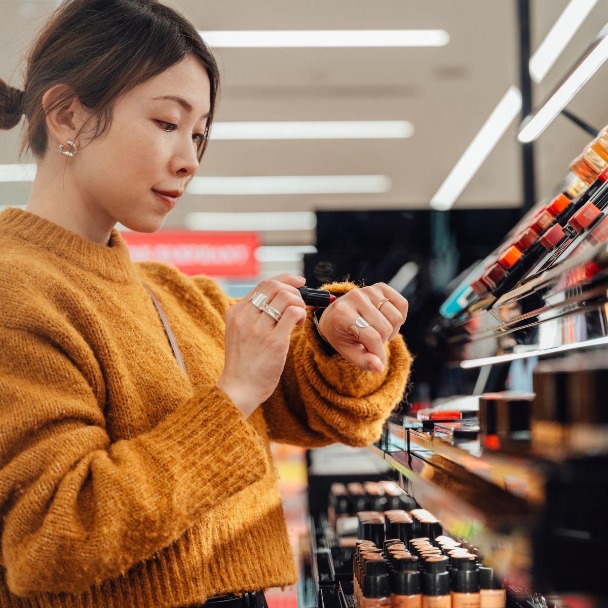 A focused woman in a knit sweater tests lipstick shade on her hand while shopping in a bright, modern cosmetics store.