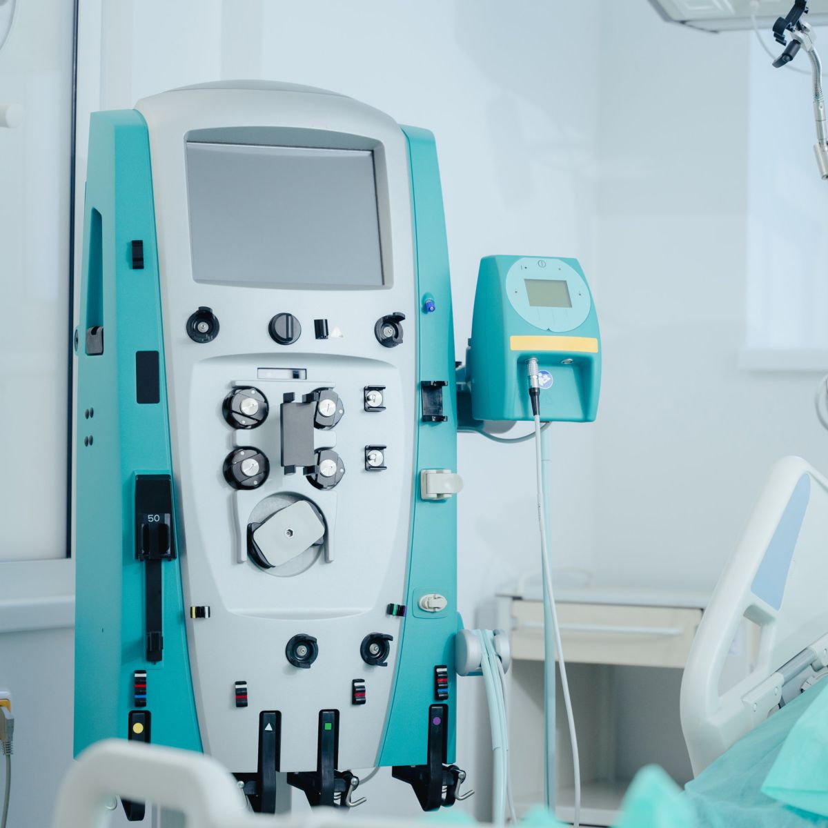A clinical dialysis machine stands next to a hospital bed with a patient monitor displaying vital signs in a sterile medical treatment room.