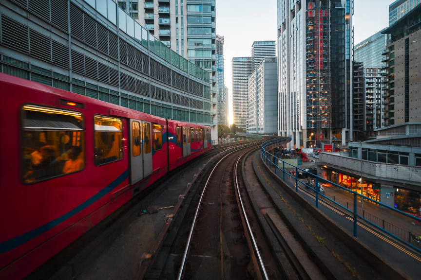 A vibrant red train travels along elevated urban tracks amidst the modern skyscrapers and bustling infrastructure of London�s financial district.