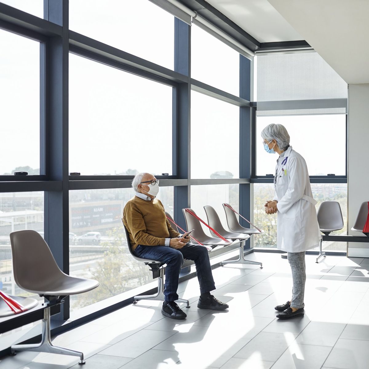 A female doctor speaks with a senior male patient wearing a face mask inside a bright, modern clinic waiting room with social distancing seating.