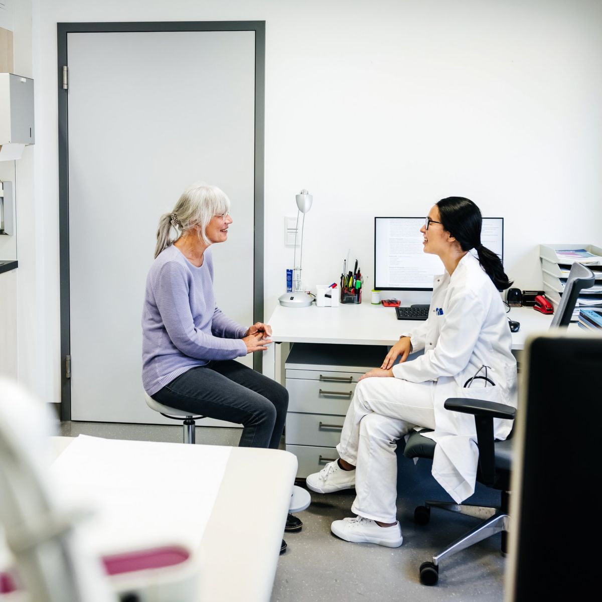 A professional female doctor engages in a friendly, supportive consultation with an elderly female patient within a modern clinic examination room.