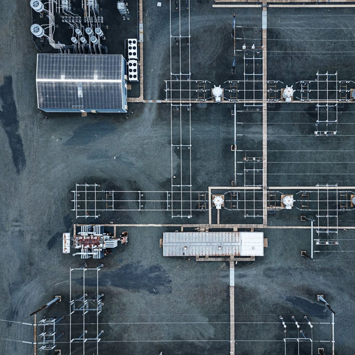A top-down aerial view captures the geometric layout of an electrical substation with power transformers and grid infrastructure on gravel terrain.