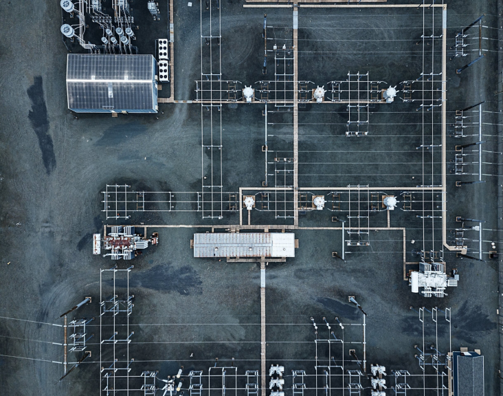 A top-down aerial view captures the geometric layout of an electrical substation with power transformers and grid infrastructure on gravel terrain.