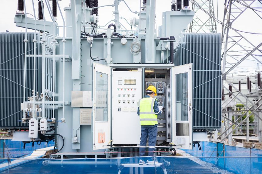 A technician in high-visibility safety gear inspects an industrial power transformer at a substation.