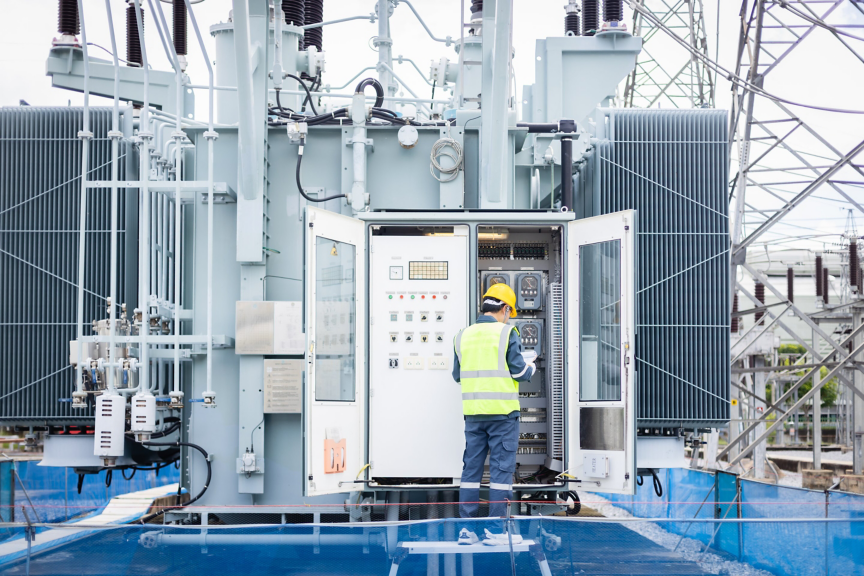 A technician in high-visibility safety gear inspects an industrial power transformer at a substation.